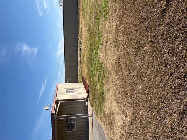 Side view of Royse City Secure Storage office building with adjacent open grass area and large metal storage unit in background under a clear blue sky.